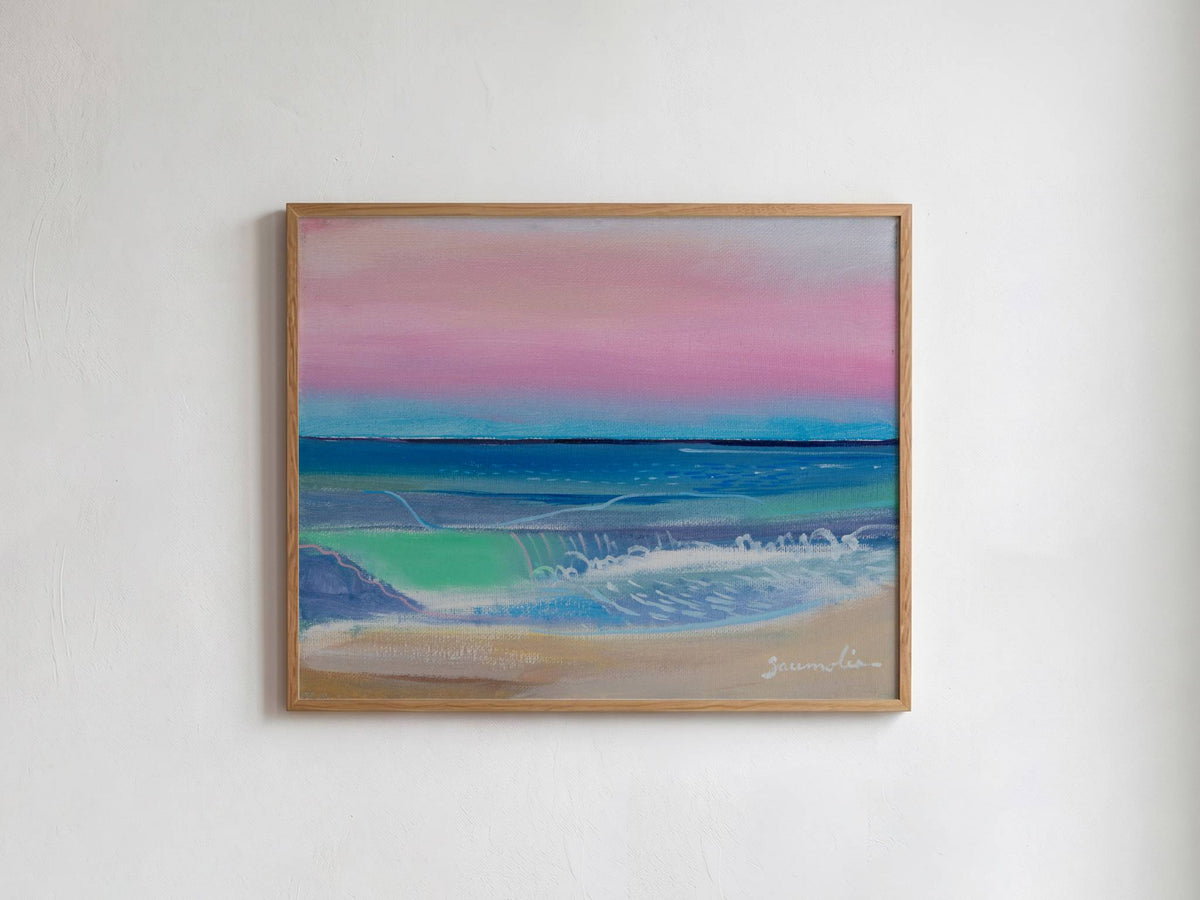 A framed landscape painting of a beach scene with pink and blue sky, turquoise water, and sandy shore, hung on a white wall.