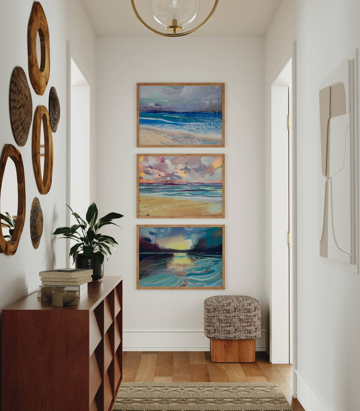A styled interior hallway featuring a vertical triptych of three framed abstract ocean paintings on the back wall, above a small upholstered stool. The hallway includes a wooden console table with books and a plant, decorative oval wall mirrors, and a glass chandelier.
