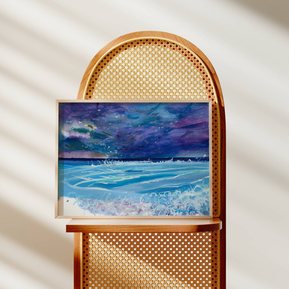 A framed painting of a stormy blue and purple ocean scene with white waves, displayed on a wooden cane-back chair against a softly lit cream wall with diagonal shadows.