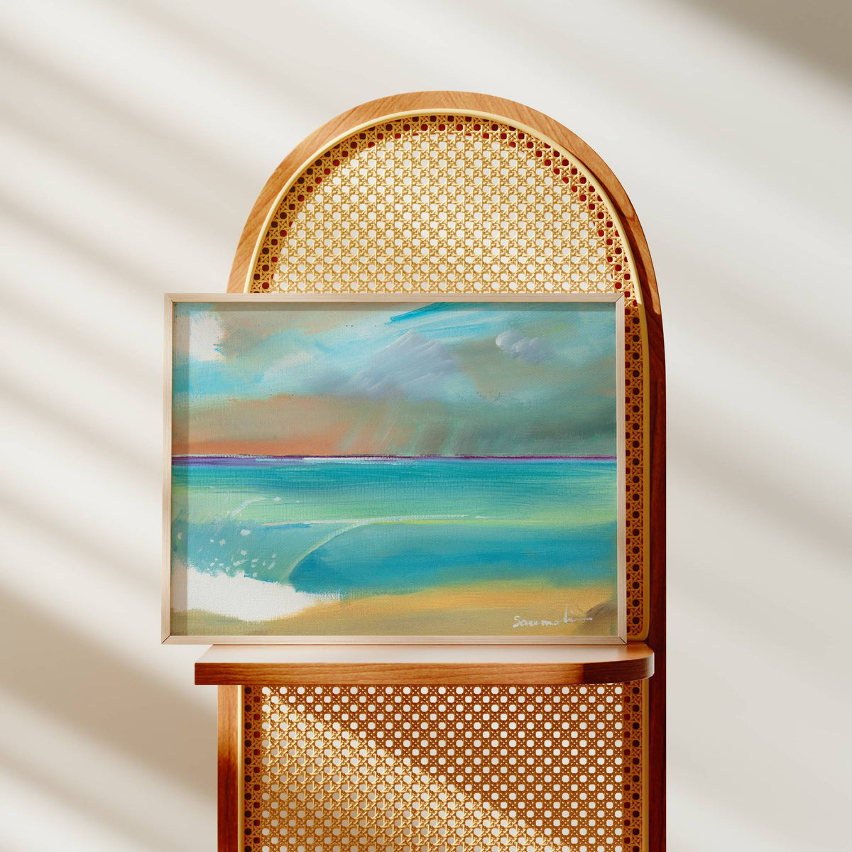 A framed pastel landscape painting of a beach and ocean under a cloudy sky, displayed on a wooden chair with a cane back in a softly lit room.