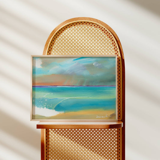 A framed pastel landscape painting of a beach and ocean under a cloudy sky, displayed on a wooden chair with a cane back in a softly lit room.