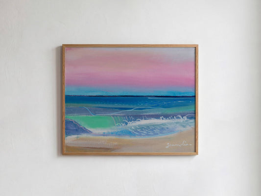 A framed landscape painting of a beach scene with pink and blue sky, turquoise water, and sandy shore, hung on a white wall.
