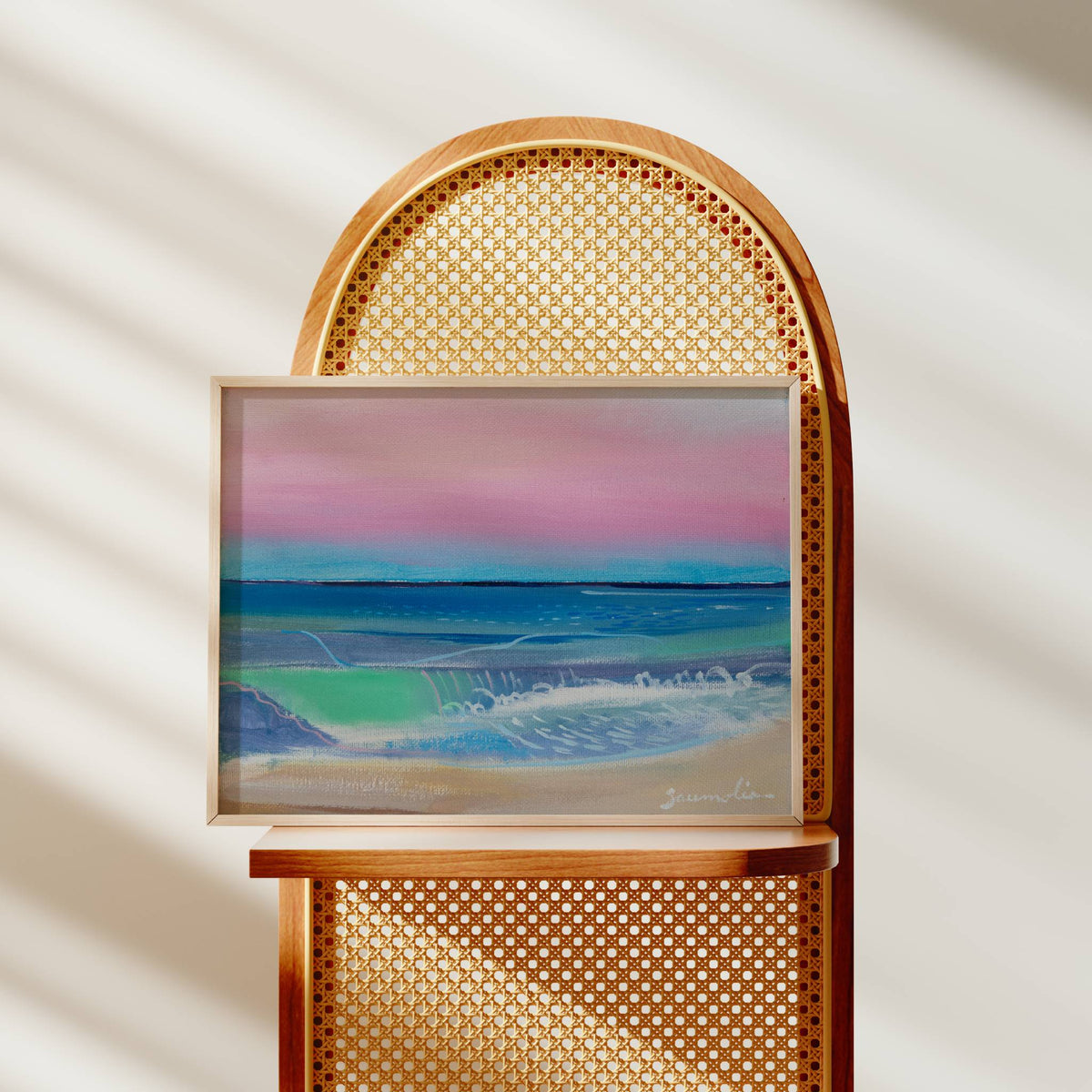 A framed landscape painting of a beach and ocean under a pink and blue sky, displayed on a wooden chair with a woven cane back against a softly lit cream wall with diagonal shadows.