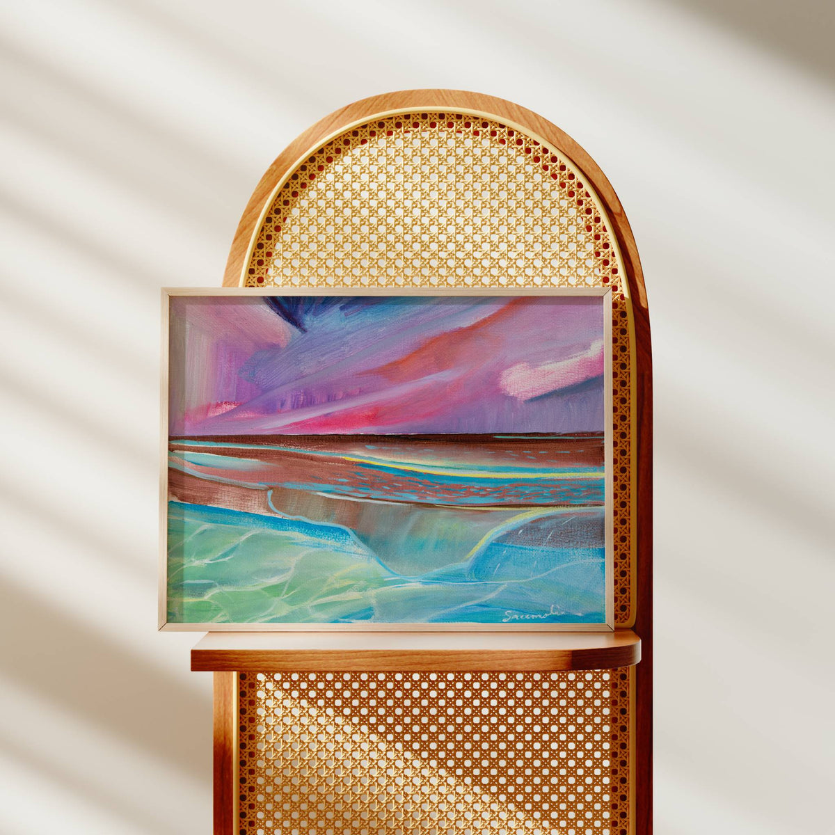 An abstract landscape painting with vibrant pink, purple, blue, and green hues, displayed in a light wood frame on a wooden cane chair against a warm beige wall with soft diagonal shadows.
