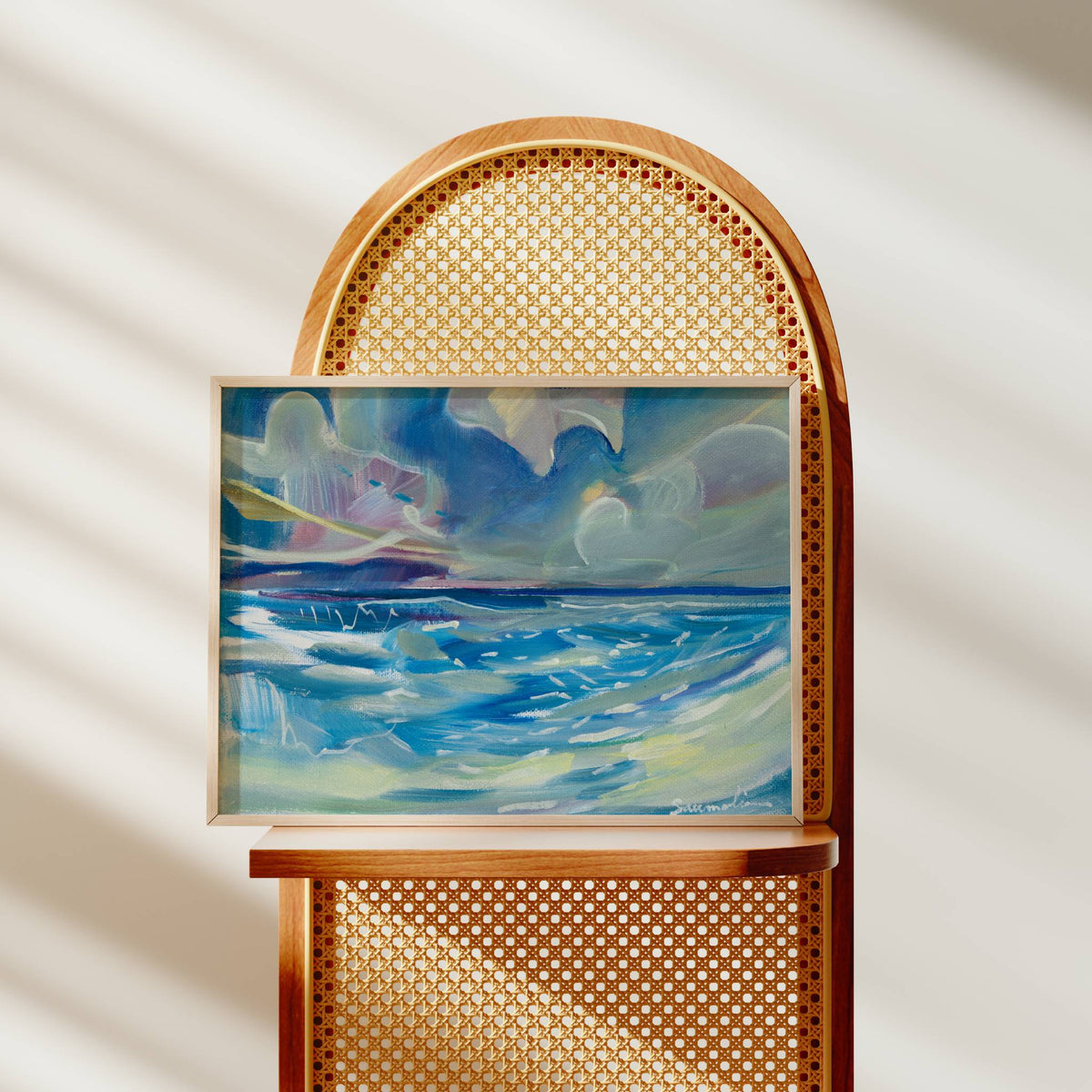 A framed abstract painting of blue ocean waves under a cloudy sky, displayed on a wooden chair with a woven cane back against a softly lit beige wall with diagonal shadows.