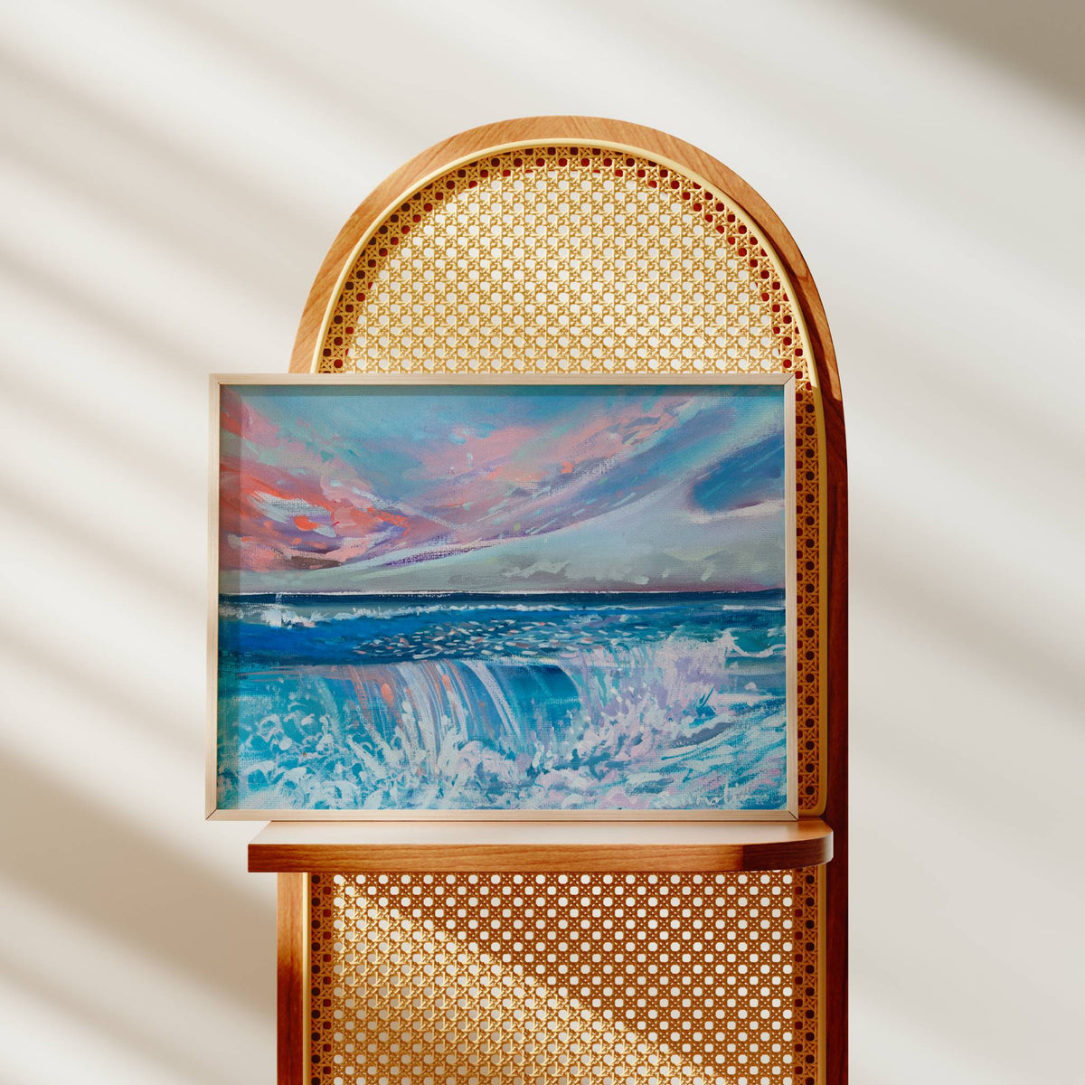 A framed abstract painting of a dramatic ocean scene with blue waves and a pink, purple, and blue sky, displayed on a wooden chair with a woven cane back against a softly lit cream wall with diagonal shadows.
