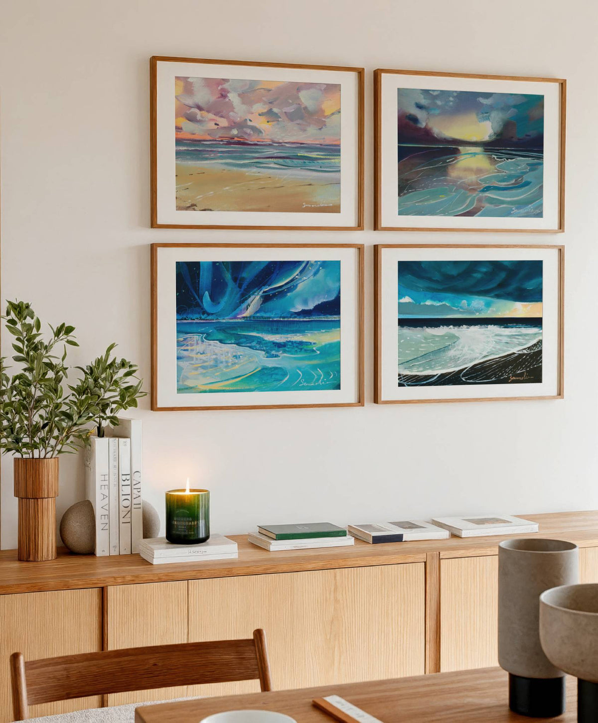 Four framed landscape paintings of coastal seas and skies arranged in a 2x2 grid on a beige wall above a light wood credenza in a styled dining room.